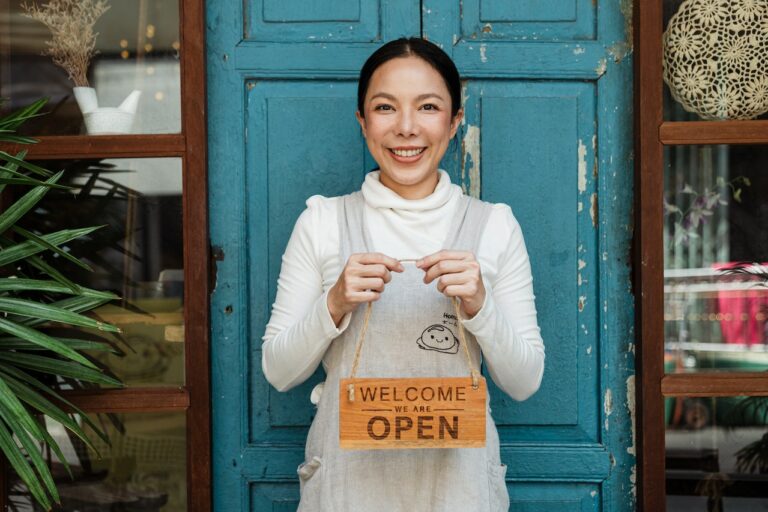 Happy business owner stands outside her shop holding a sign that says "welcome we are open". Learn more about startup and licensing services to get your business started.
